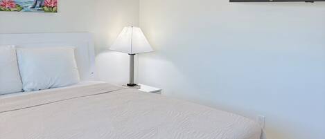 Un dormitorio con cama, televisor montado en la pared, mesita de noche con lámpara y un cuadro de flamencos.