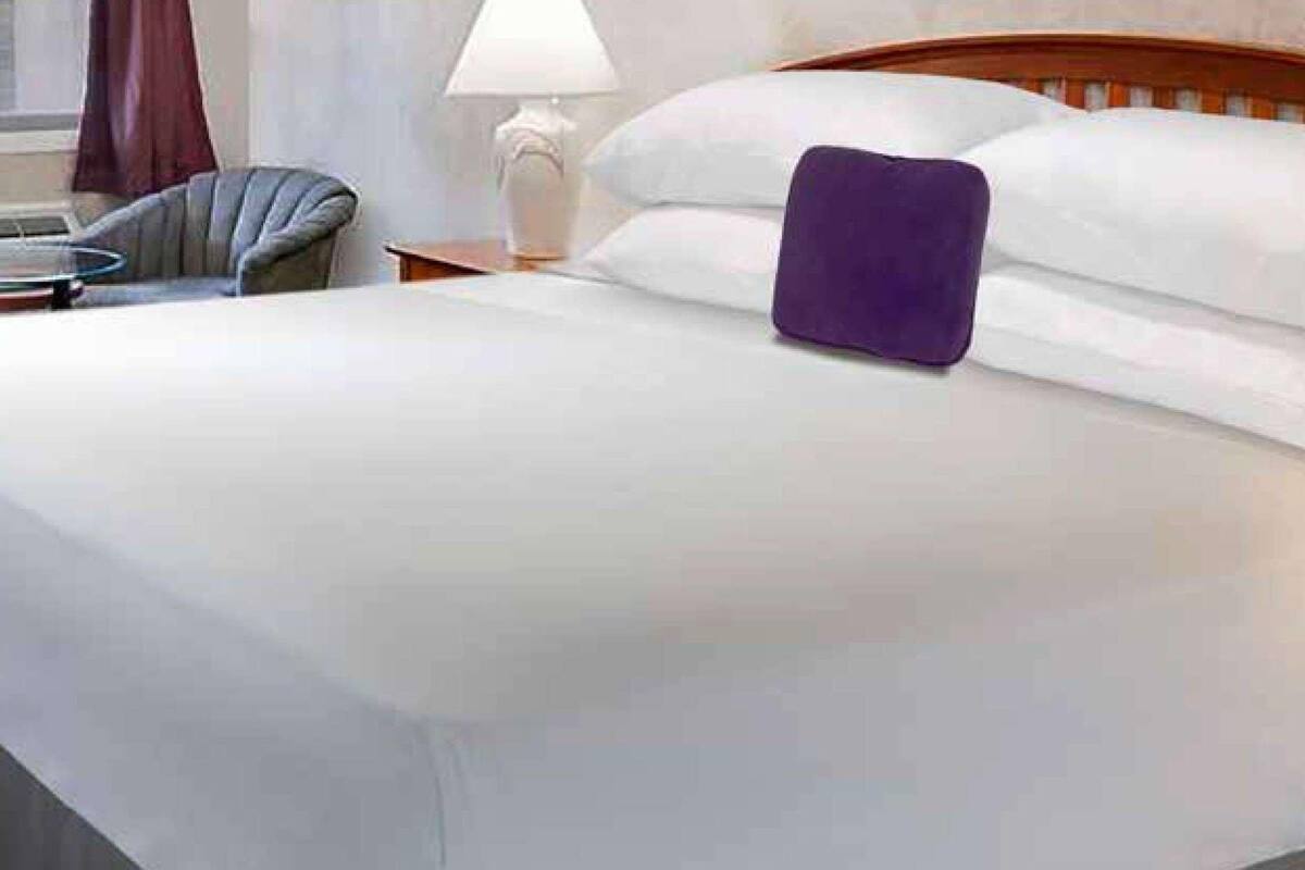 Een net opgemaakt bed met witte lakens en een paars kussen, een houten hoofdbord en een nachtkastje met een lamp.