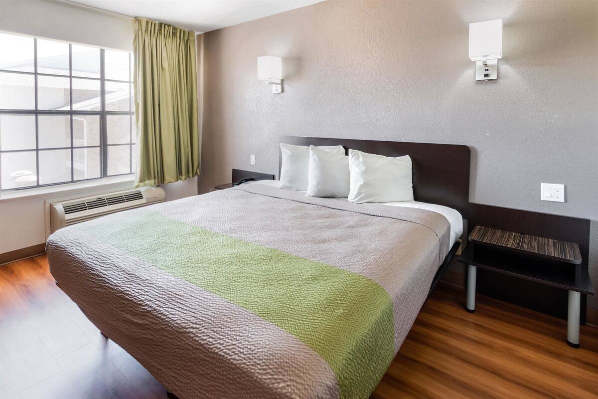 A hotel room with a large bed, a bedside table, a wall-mounted lamp, and a window with curtains.