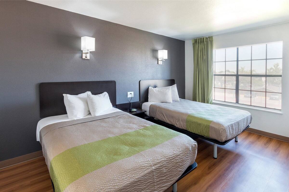 A hotel room with two beds, a large window with curtains, and wall-mounted lights.
