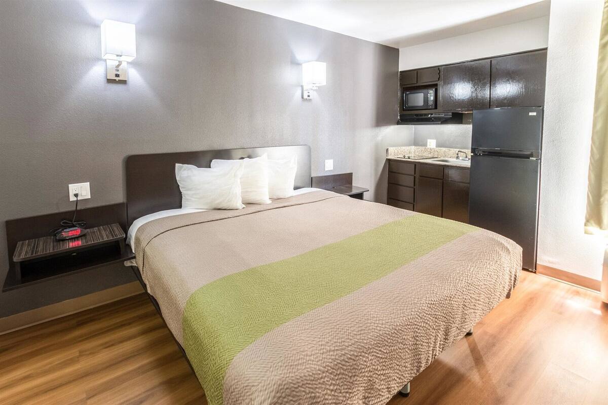 A compact hotel room with a bed, bedside table, wall-mounted lamp, and a kitchenette with a microwave and refrigerator.