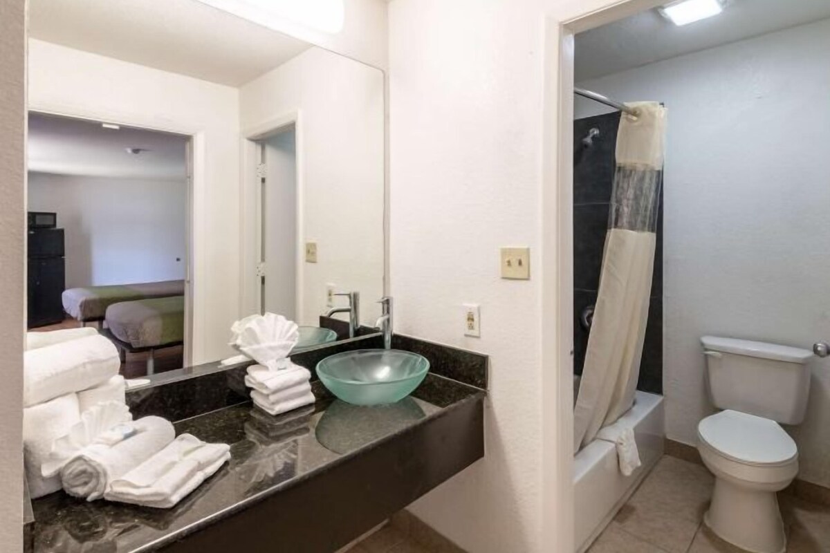 Un baño con un lavamanos de vidrio, un inodoro, una cortina de ducha y un espejo que refleja una cama y un televisor.