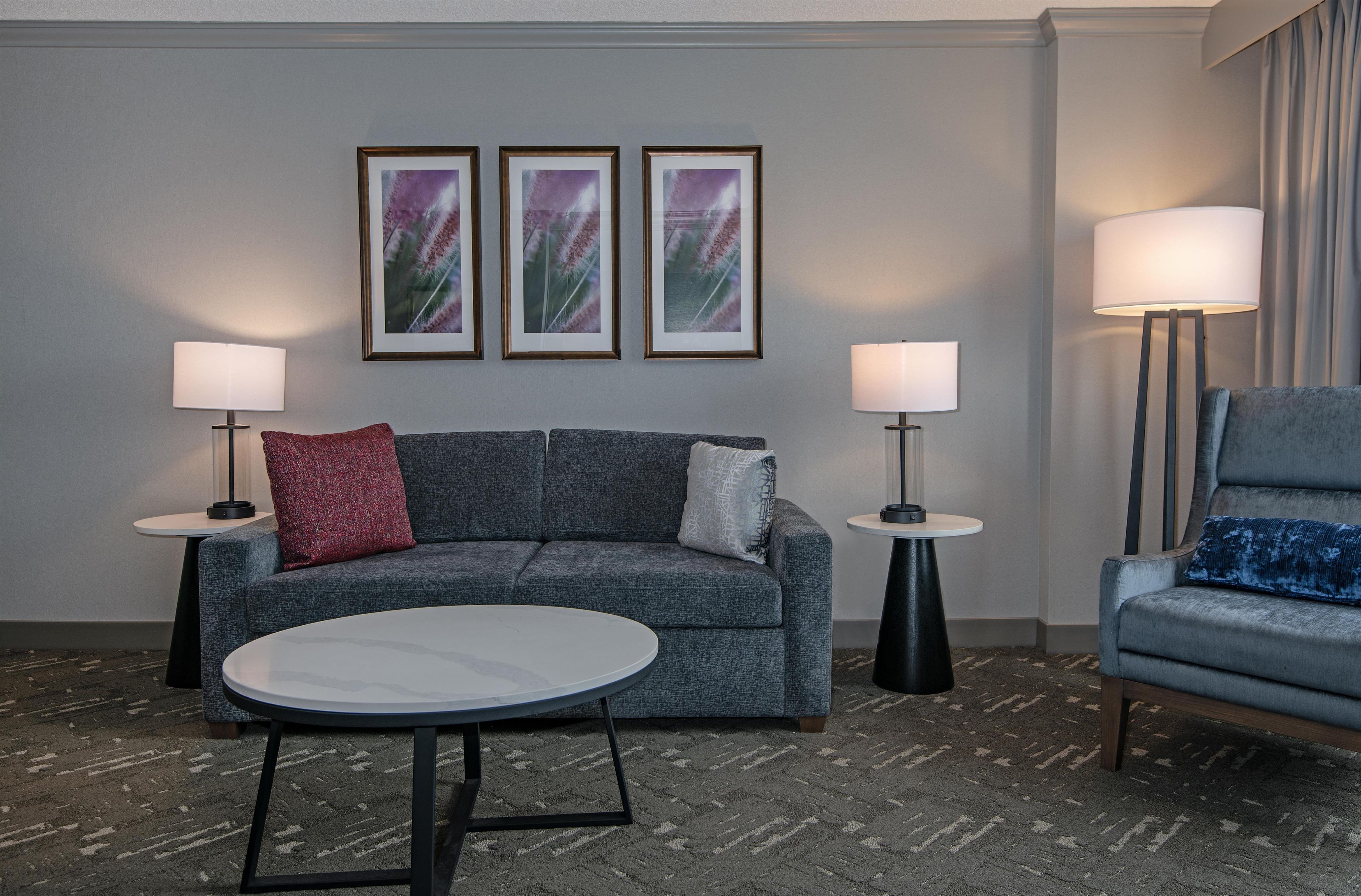 Una habitación de hotel moderna con un sofá gris, una mesa redonda, dos mesitas de noche con lámparas y cuadros en la pared.