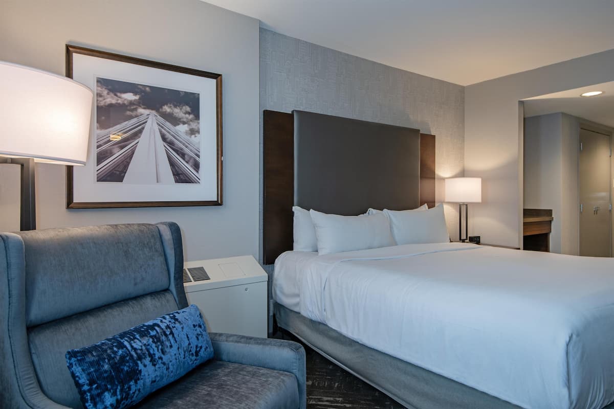 A hotel room with a bed, a chair, a desk, a lamp, and a framed picture on the wall.