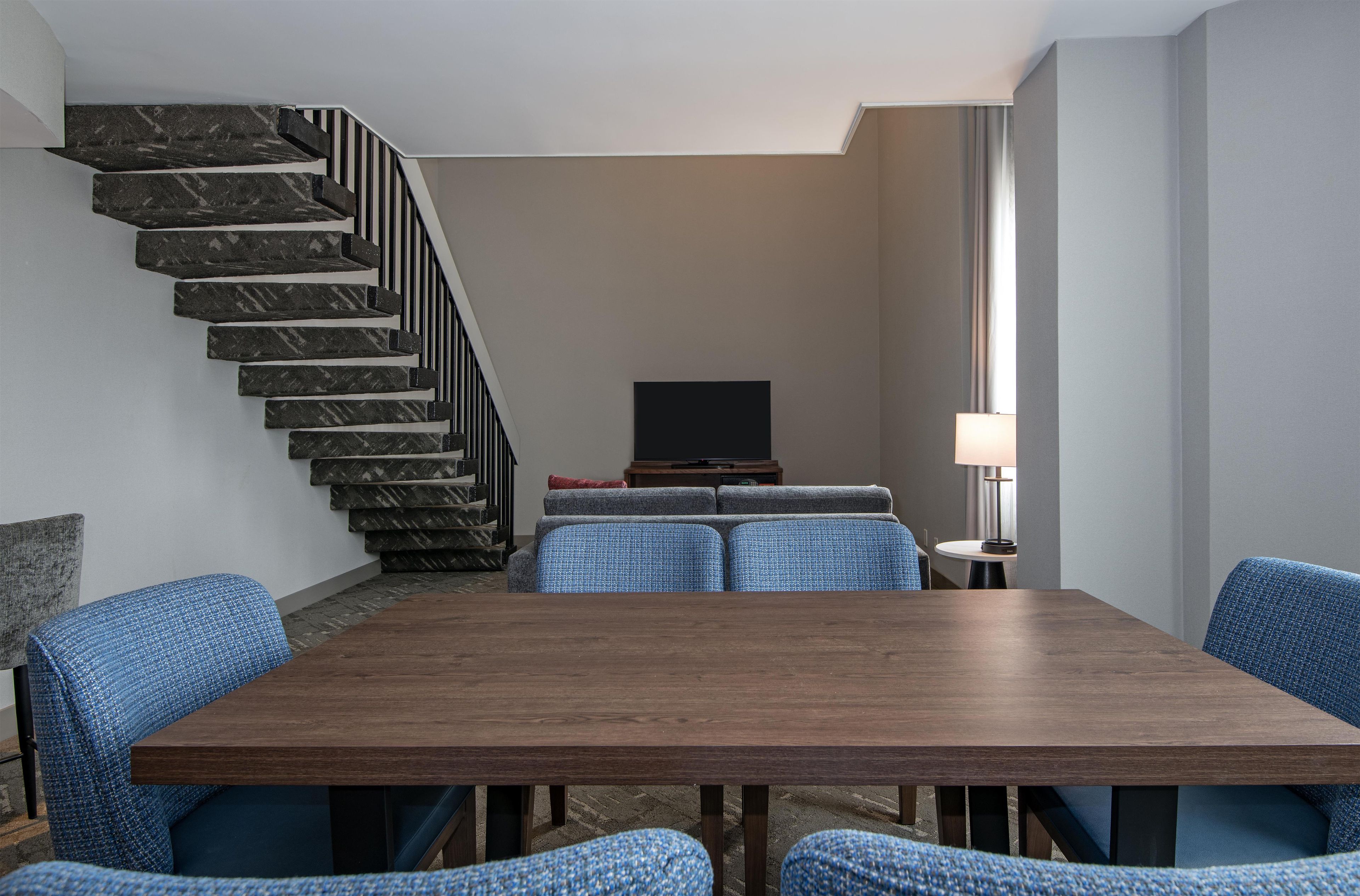 Una sala de estar moderna con una mesa de comedor, sillas tapizadas en azul, un conjunto de sofás, un televisor y una escalera.