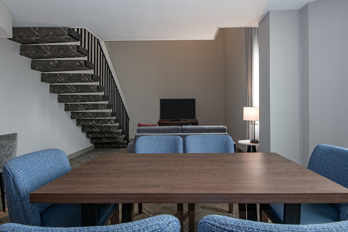 A modern living room with a dining table, blue upholstered chairs, a sofa set, a TV, and a staircase.