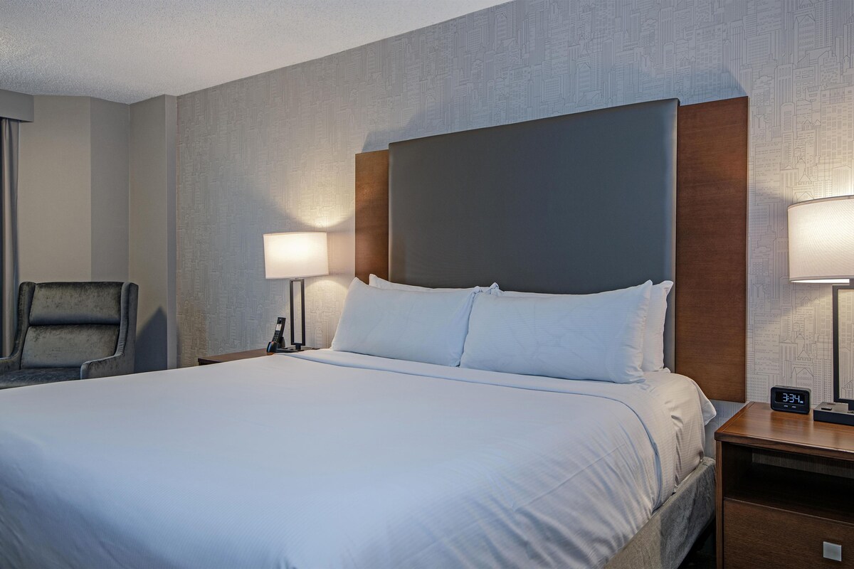 A hotel room with a large bed, two bedside lamps, a chair, and a nightstand with a clock.