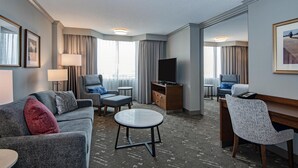 Habitación de hotel con sofá, sillón, mesa redonda, televisor y escritorio con silla.