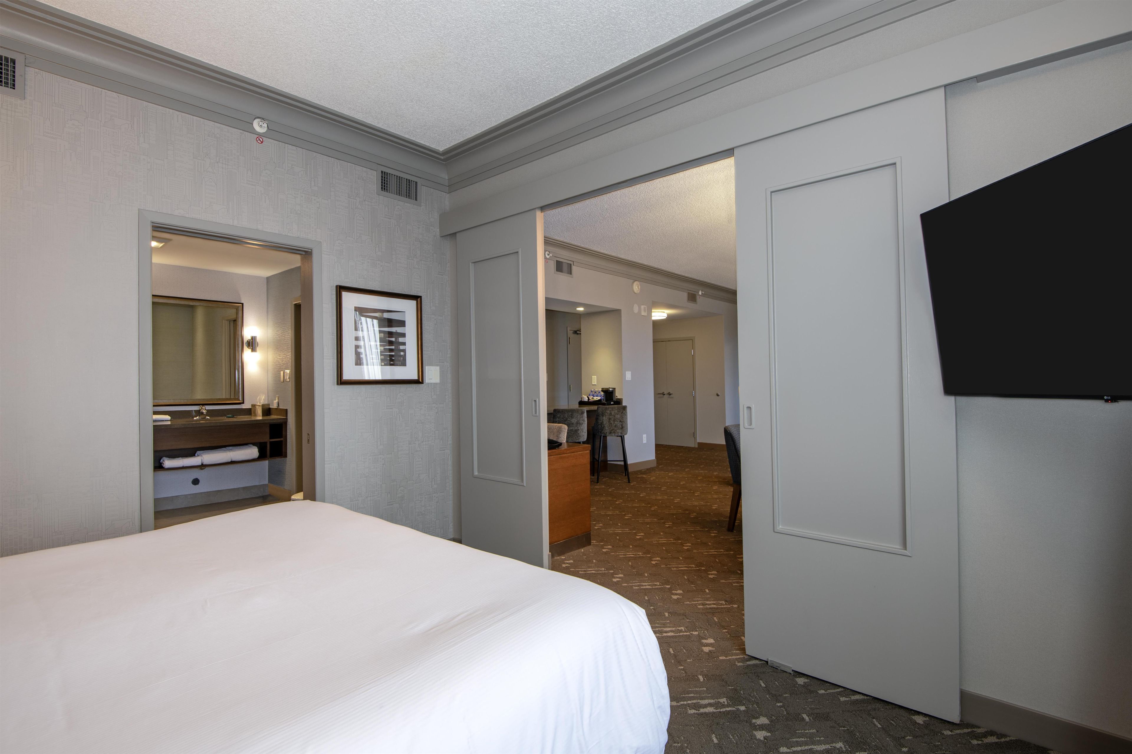 Habitación de hotel con una cama grande, baño con espejo y perchero, televisión en la pared y pasillo que conduce a otras habitaciones.