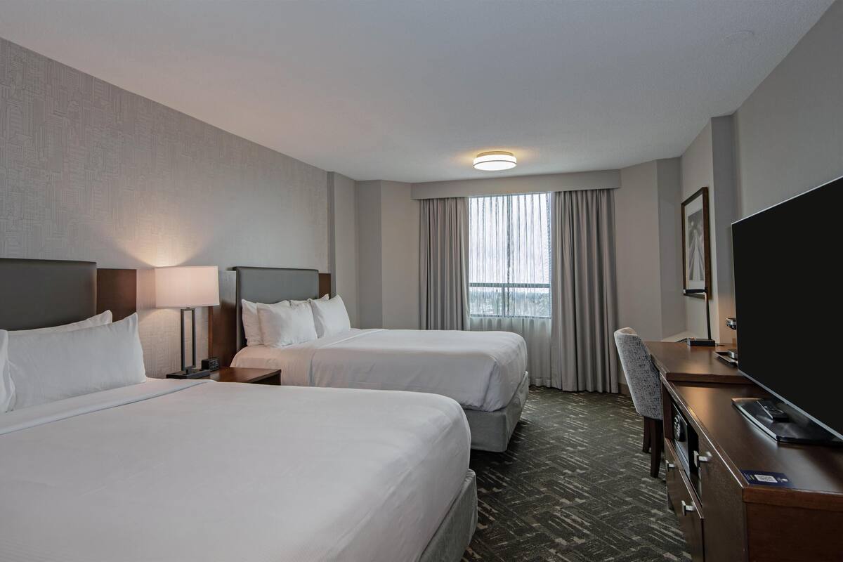 A hotel room with two beds, a desk, a chair, a TV, and a window with curtains.