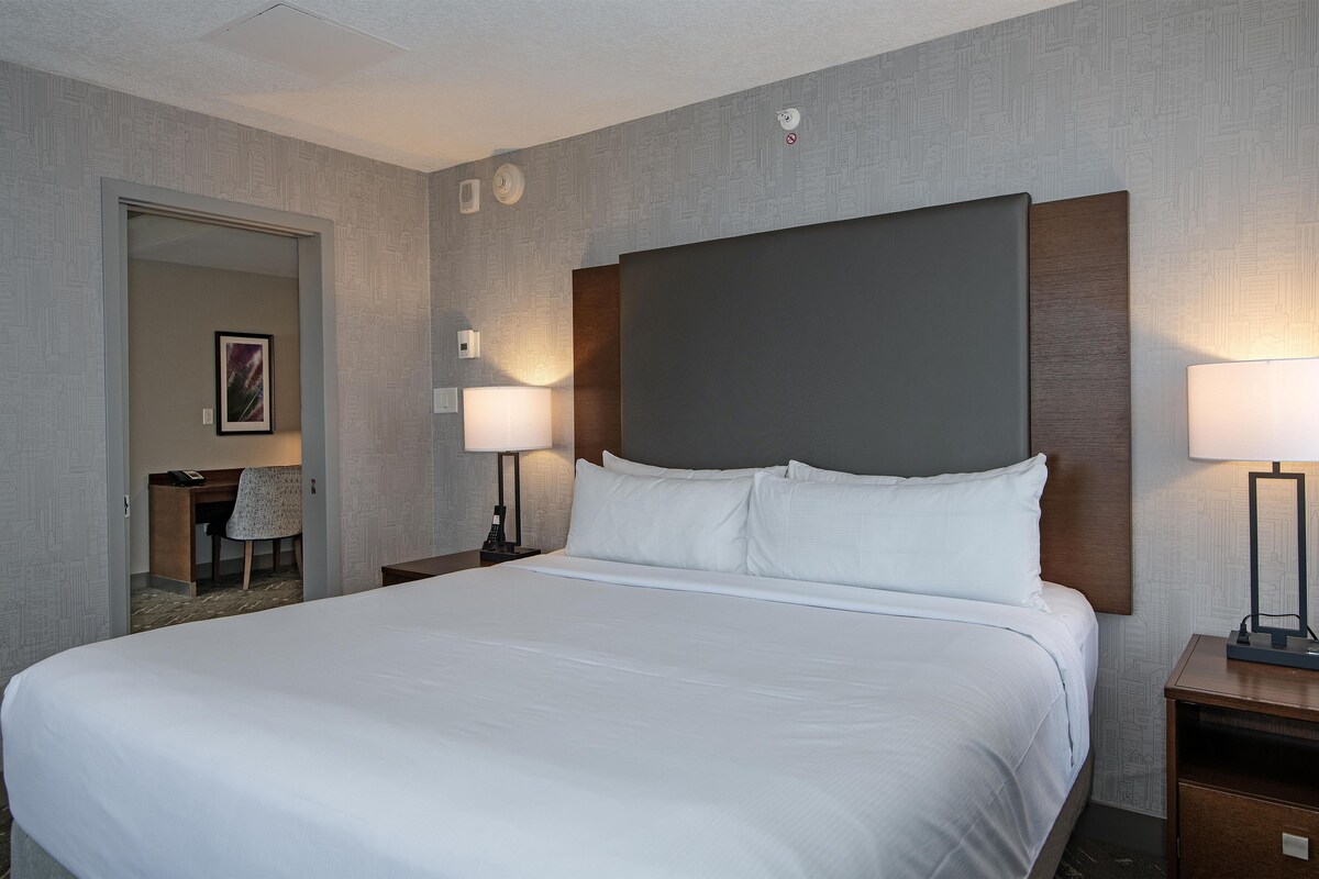 A hotel room with a large bed, two bedside tables with lamps, a desk with a chair, and a door leading to another room.