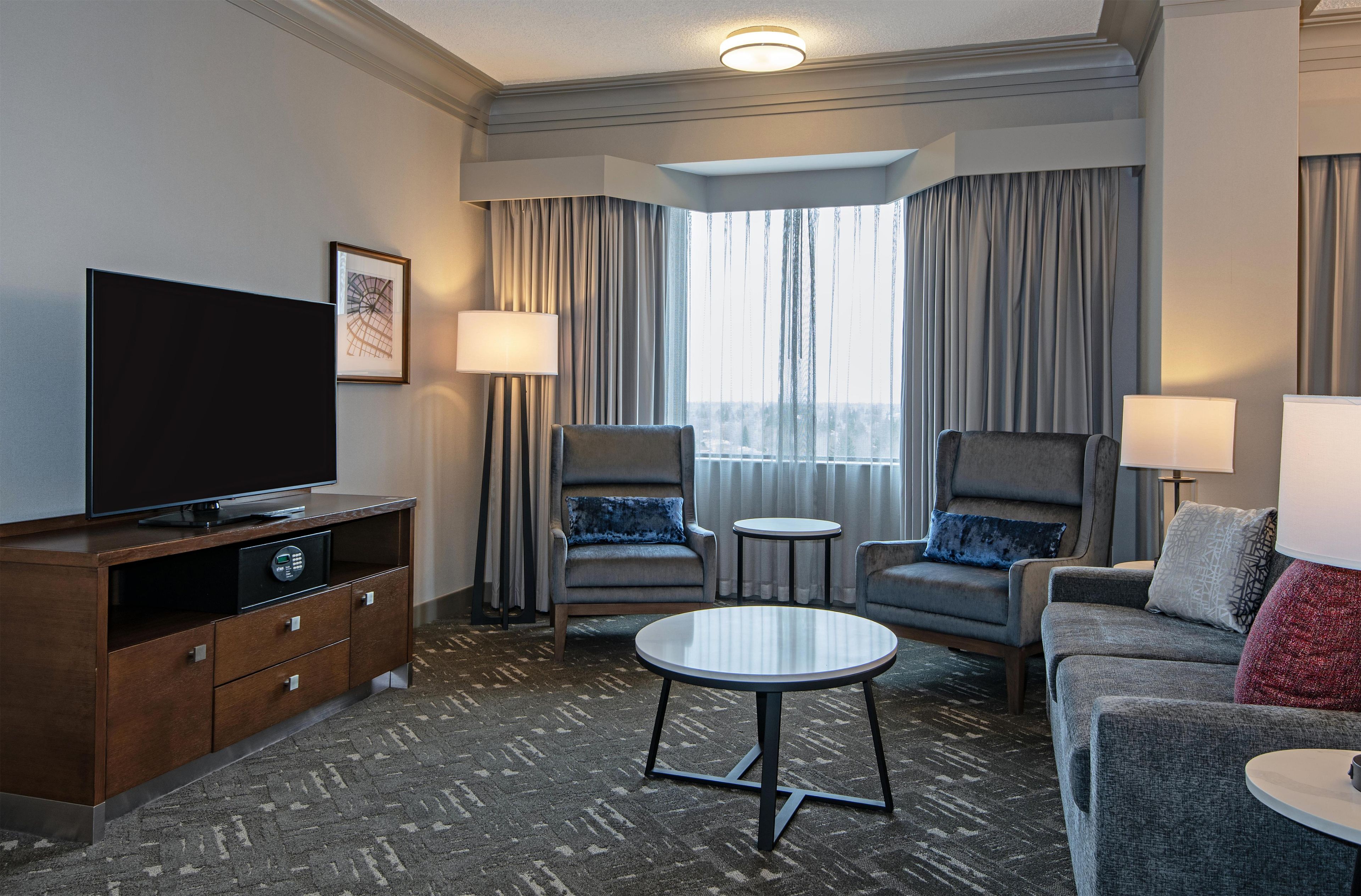 A hotel room with a flat-screen TV, two armchairs, a sofa, a small table, a cabinet, and a window with curtains.