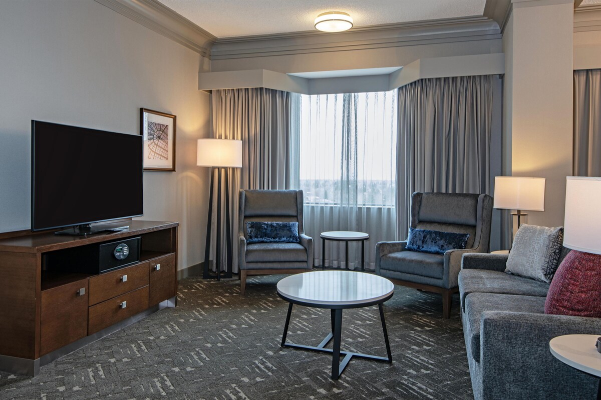 A hotel room with a flat-screen TV, two armchairs, a sofa, a small table, a cabinet, and a window with curtains.