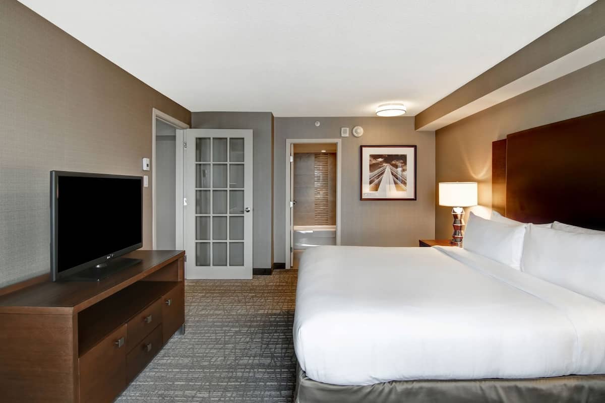 A hotel room with a bed, a TV, a desk, a lamp, and a door leading to a bathroom.