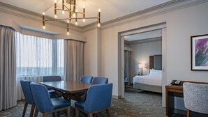 Una habitación de hotel con comedor, cama, escritorio y un amplio ventanal con cortinas.