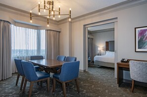 A hotel room with a dining area, a bed, a desk, and a large window with curtains.