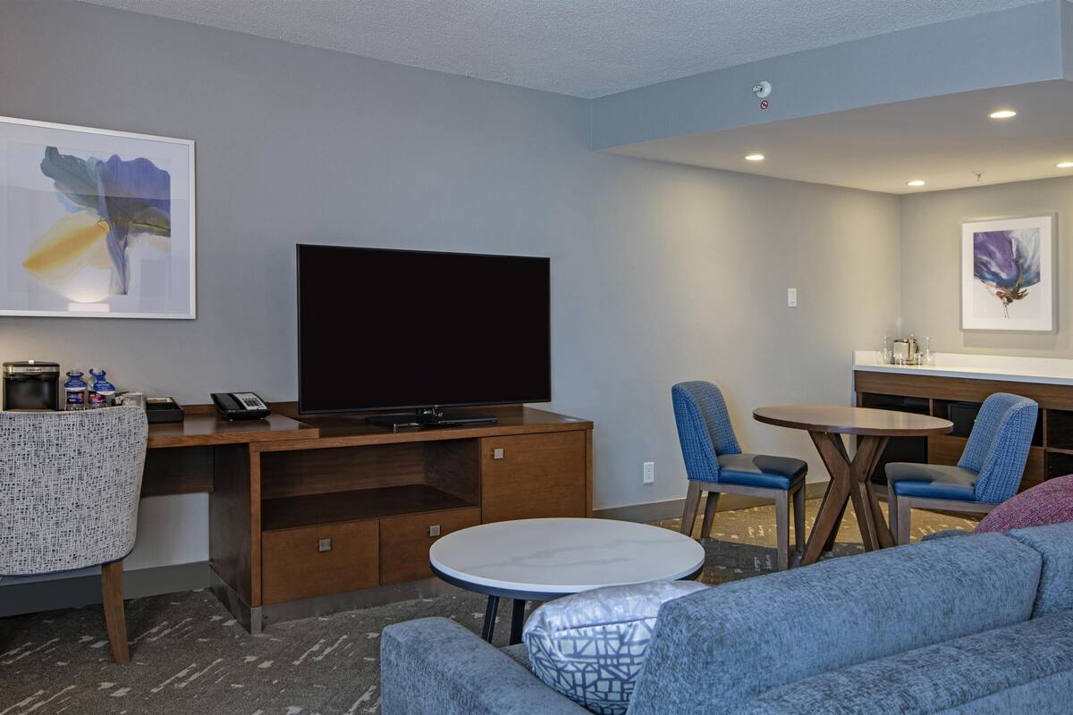 A modern hotel room with a flat-screen TV, a dining area with blue chairs, a round table, and a sofa.