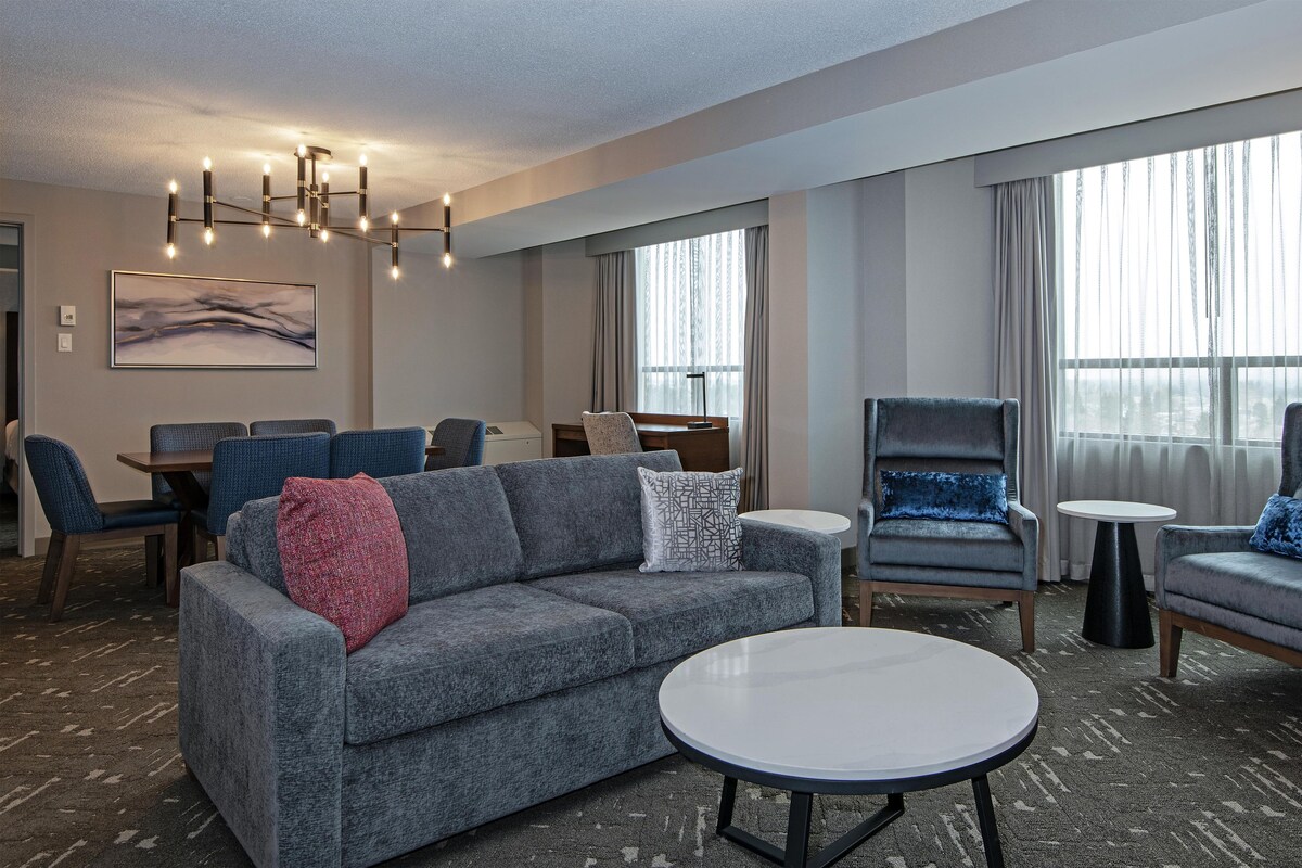 A modern hotel room with a grey sofa, round coffee table, and matching armchairs. There is a dining area with a table and chairs, and a large window with curtains.