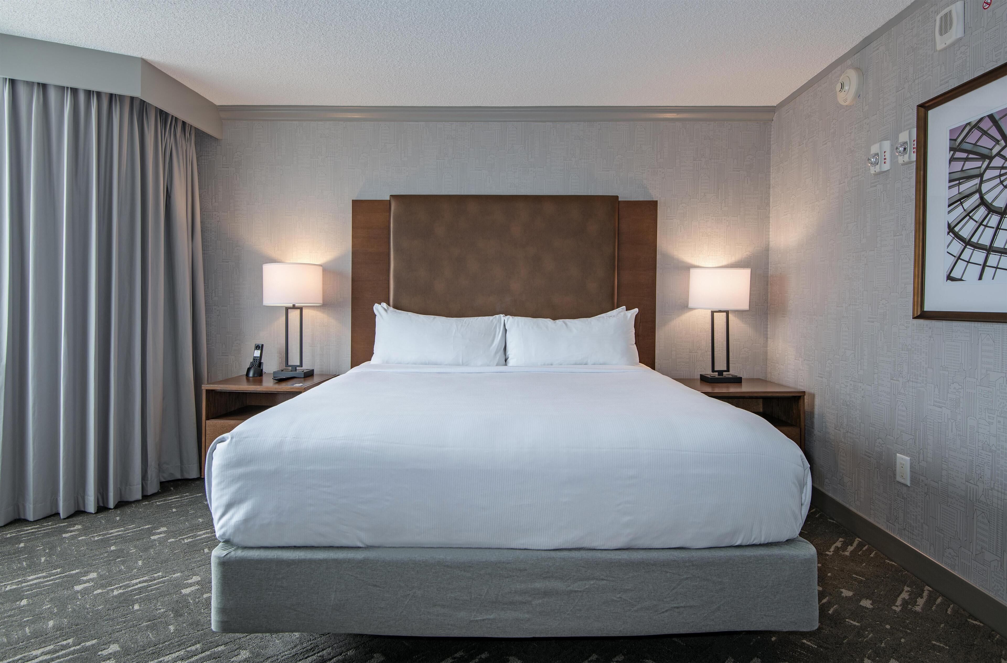 A hotel room with a large bed, two bedside tables, a headboard, a wall-mounted artwork, and a window with curtains.