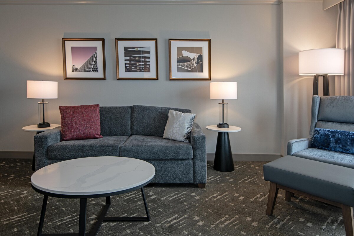 A modern hotel room with a grey sofa, two side tables with lamps, a round coffee table, and framed artwork on the wall.