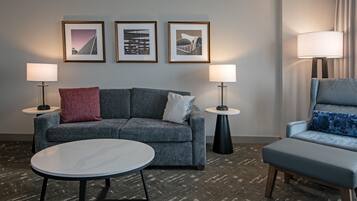 Habitació d'hotel moderna amb un sofà gris, dues taules laterals amb làmpades, una taula de centre rodona i obra d'art emmarcada a la paret.