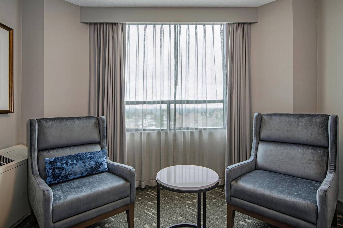 A hotel room with two grey armchairs, a small round table, and a large window with curtains.