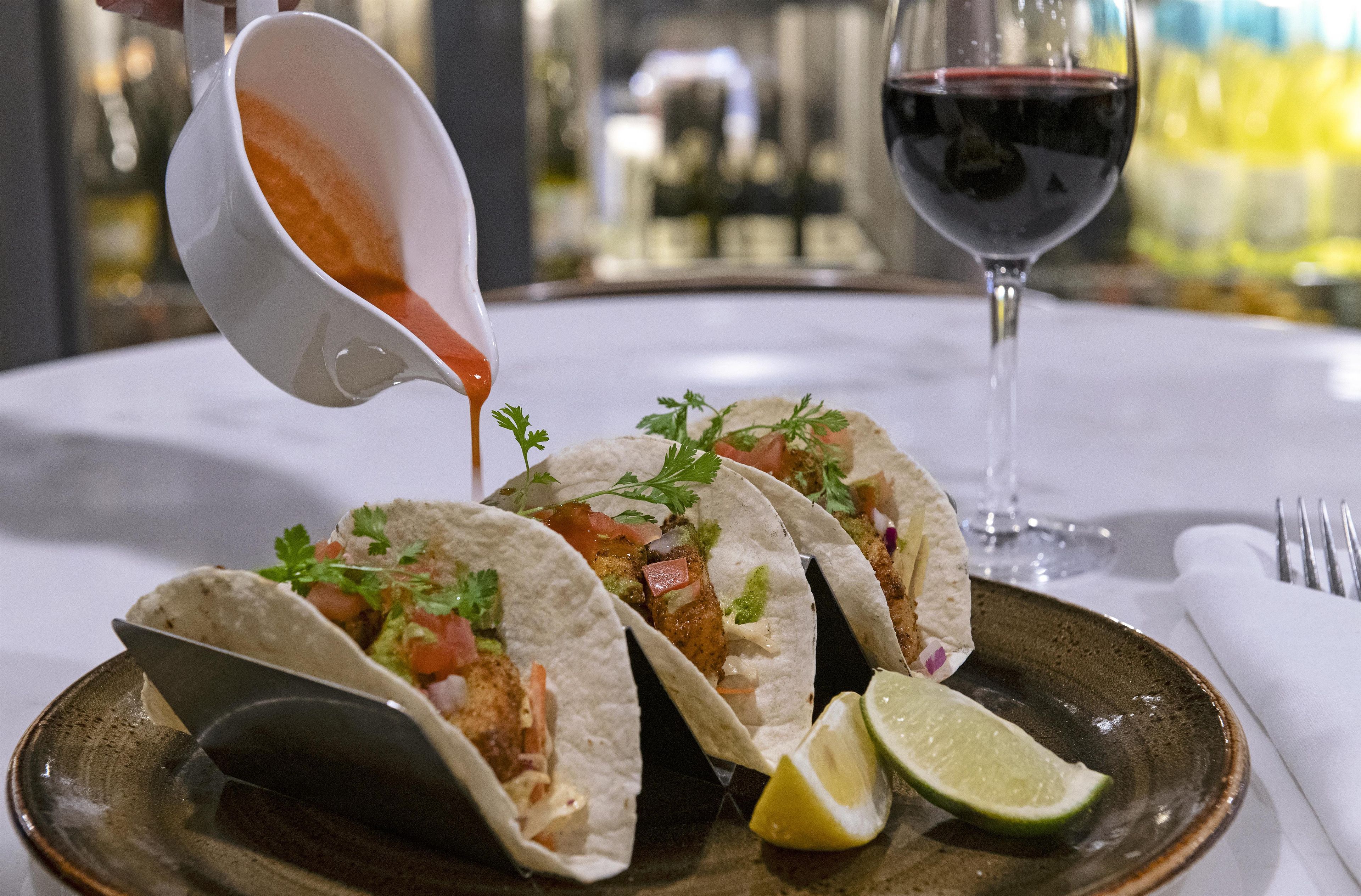 Three tacos on a plate with a lime wedge, a glass of red wine, and a white sauce being poured.