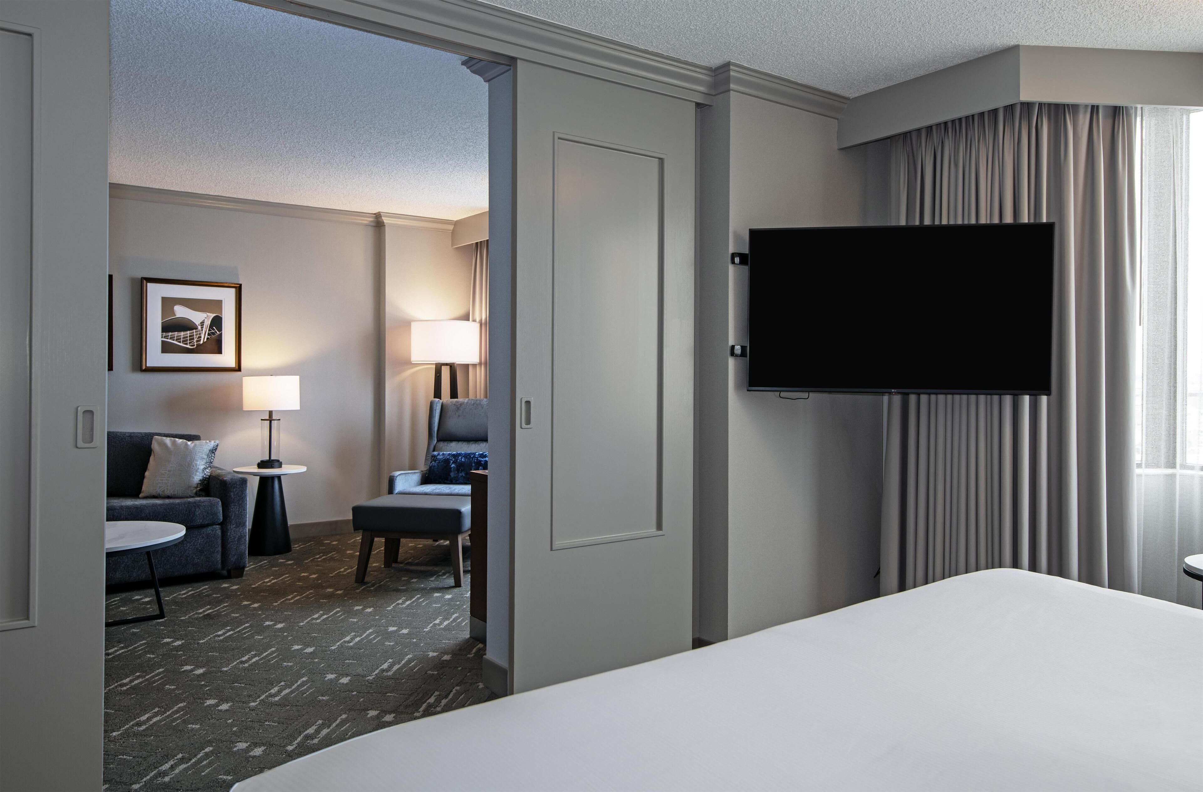 A hotel room with a bed, a TV mounted on the wall, a desk, a chair, a lamp, and a framed picture on the wall.