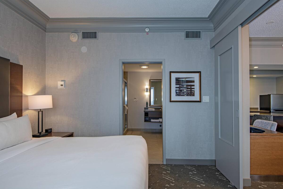 A hotel room with a bed, a nightstand, a lamp, a framed picture on the wall, and a bathroom visible through an open door.