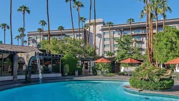 Relax by the resort pool surrounded by lush greenery and palm trees, with a waterfall feature and comfortable outdoor seating.