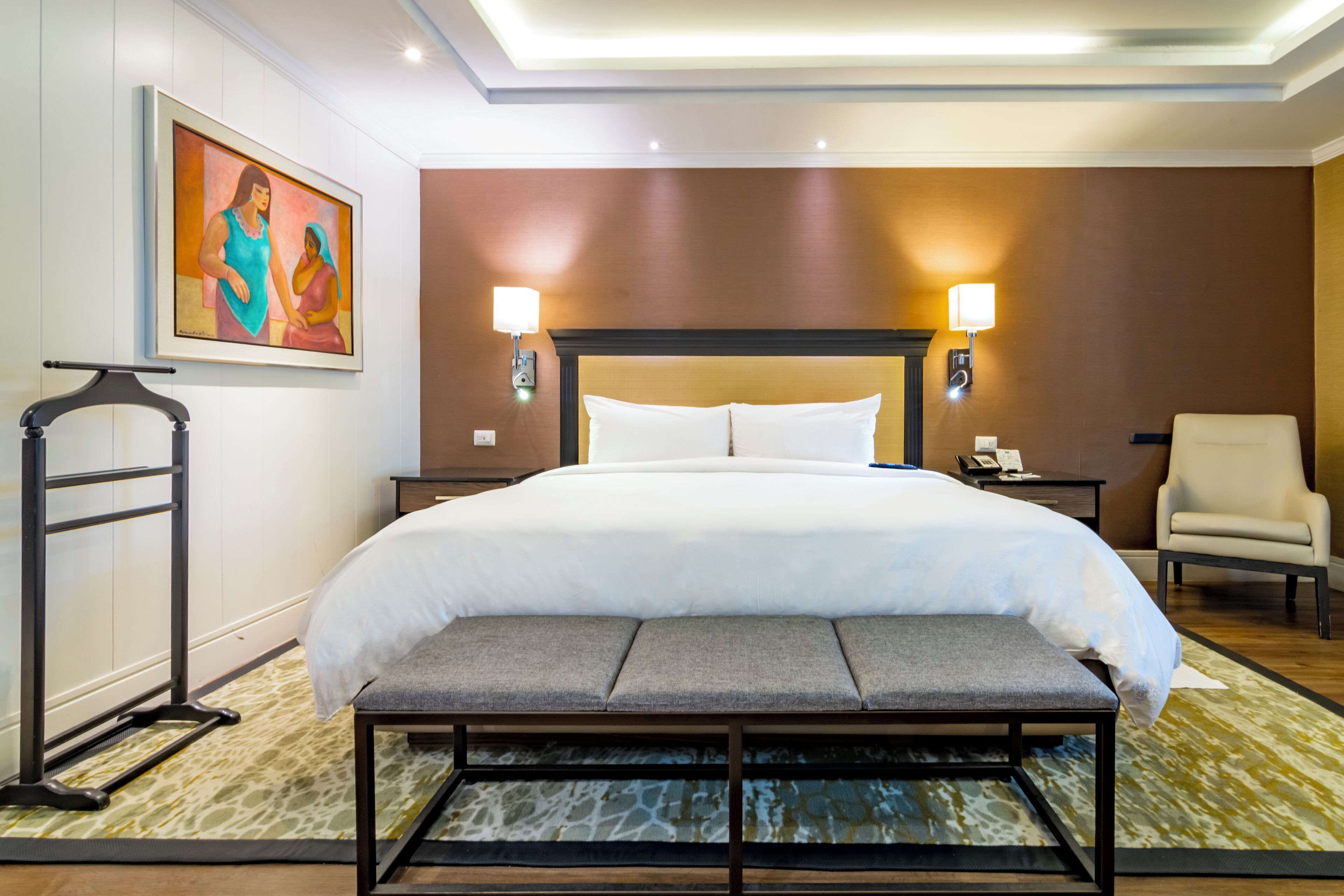 A hotel room with a large bed, bedside lamps, a bench, a chair, and a painting on the wall.