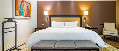 A hotel room with a large bed, bedside lamps, a bench, a chair, and a painting on the wall.
