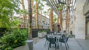 An outdoor patio area with tables and chairs, surrounded by palm trees and potted plants. - Hotel Lucerna Mexicali (Mexicali)