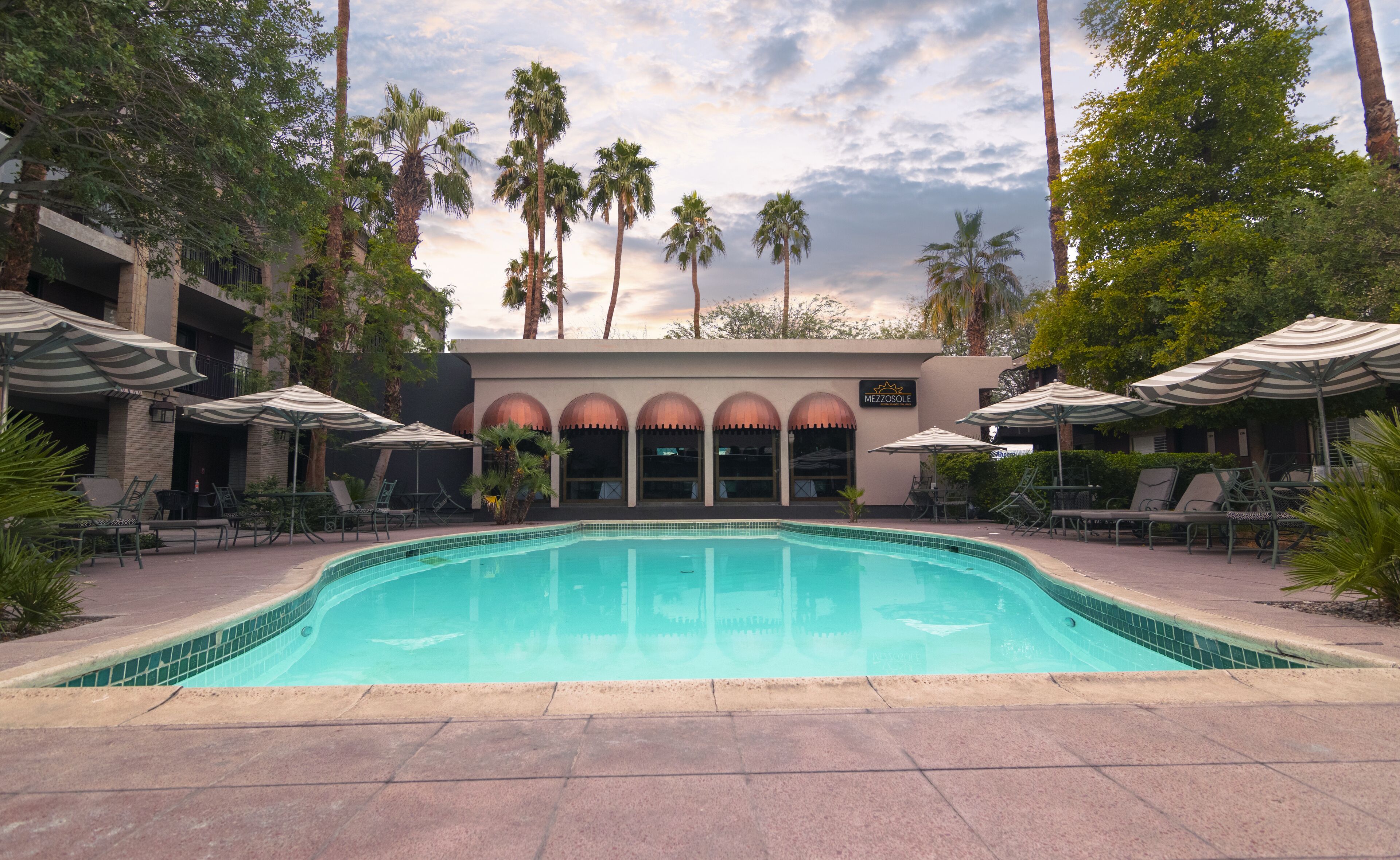 A swimming pool with lounge chairs and umbrellas, surrounded by palm trees and a building with arched windows.