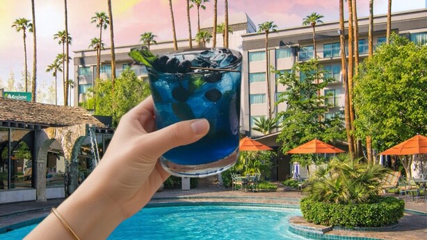 A hand holding a blue drink with ice cubes, a swimming pool, palm trees, and a hotel building in the background. - Hotel Lucerna Mexicali (Mexicali)
