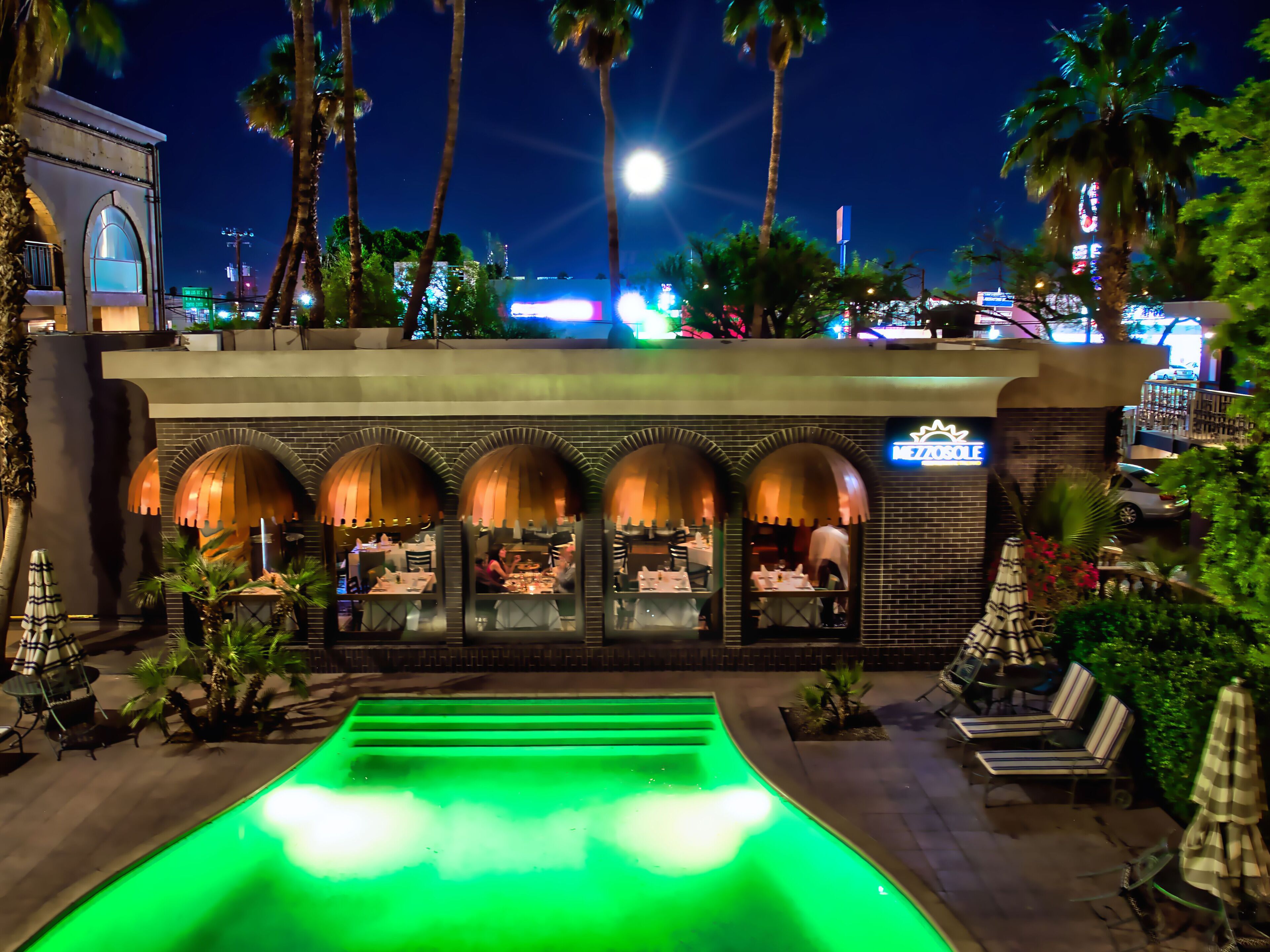A restaurant with outdoor seating, a green-lit pool, and palm trees.