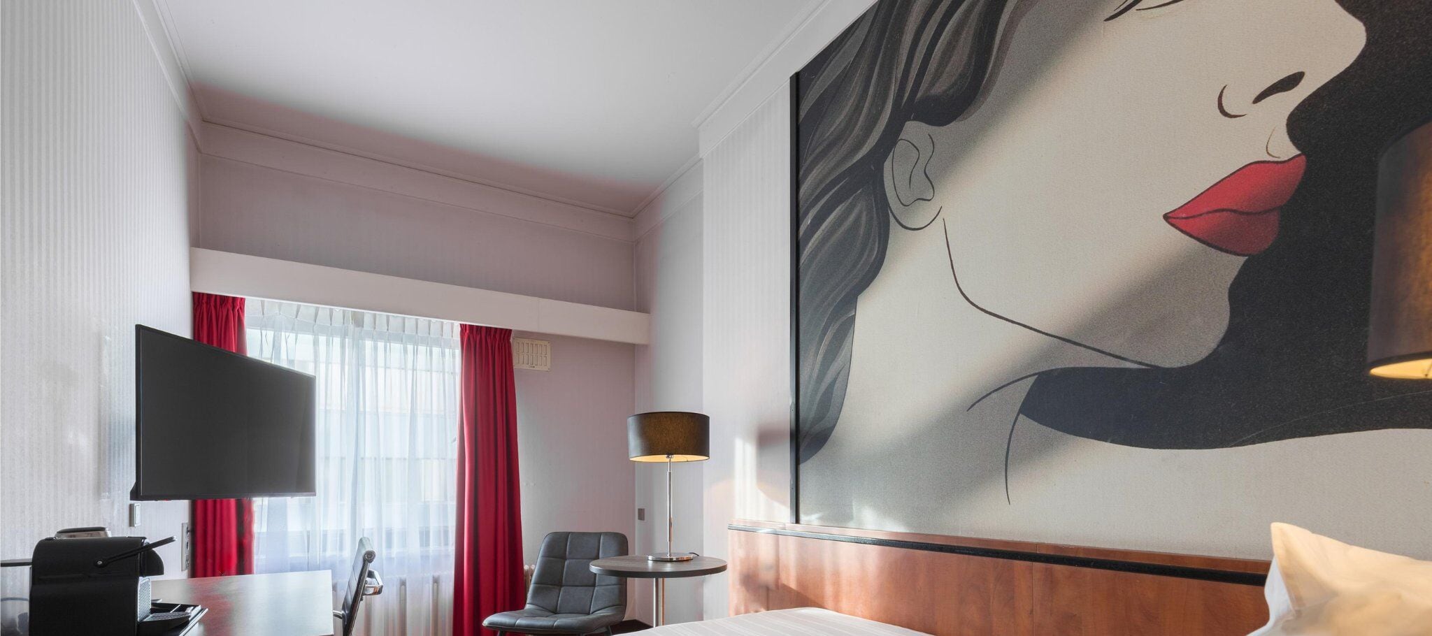 A hotel room with a bed, a desk, a chair, a TV, a lamp, and a large painting on the wall.