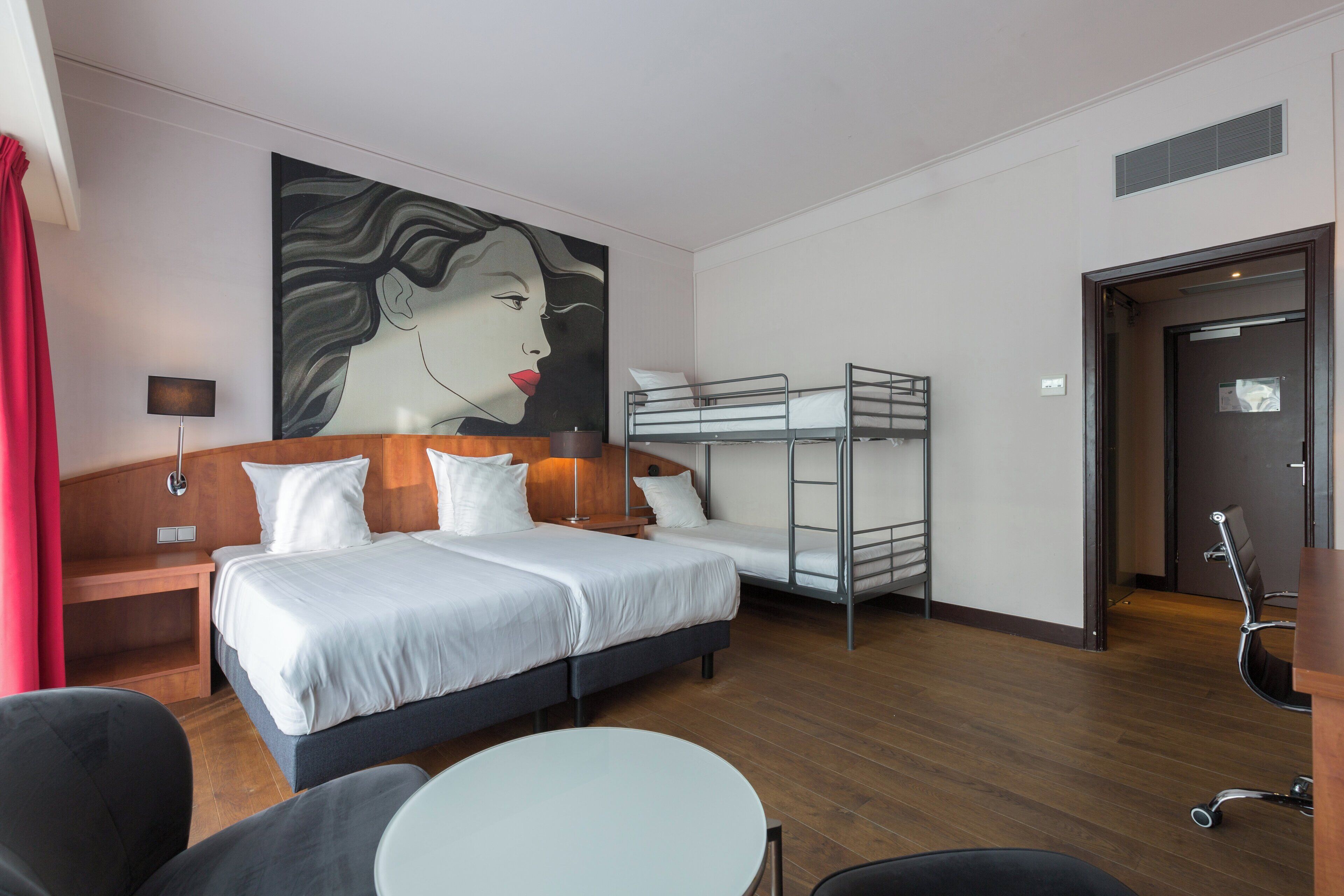 A hotel room with a bed, a desk, a chair, a bunk bed, and a painting on the wall.
