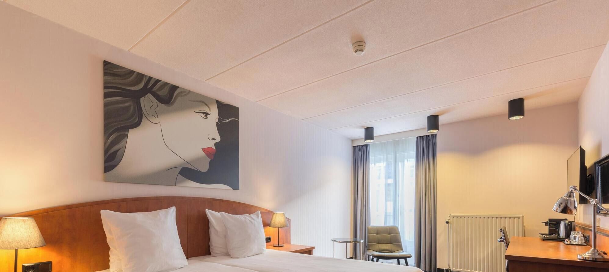 Ein Hotelzimmer mit einem Bett, einem Schreibtisch mit Lampe, einem Sessel, einem Heizkörper und einem großen Fenster mit Vorhängen.