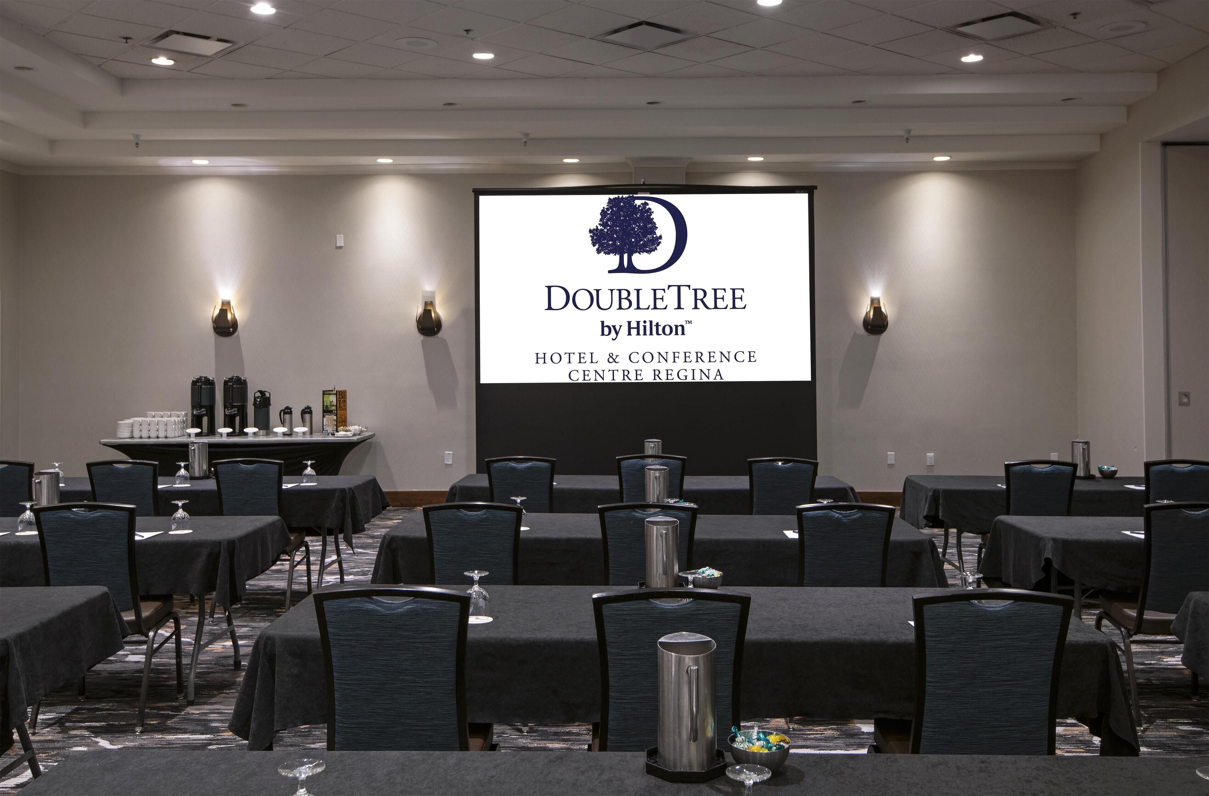 Un salón de conferencias con mesas, sillas, un atril y una pantalla mostrando el logotipo de DoubleTree by Hilton.