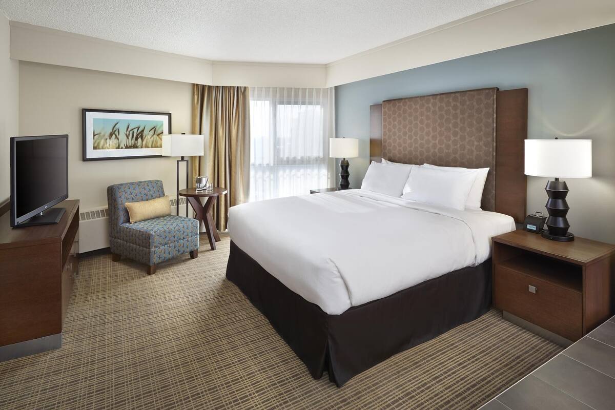 A hotel room with a large bed, a TV, a desk, a chair, and a painting on the wall.