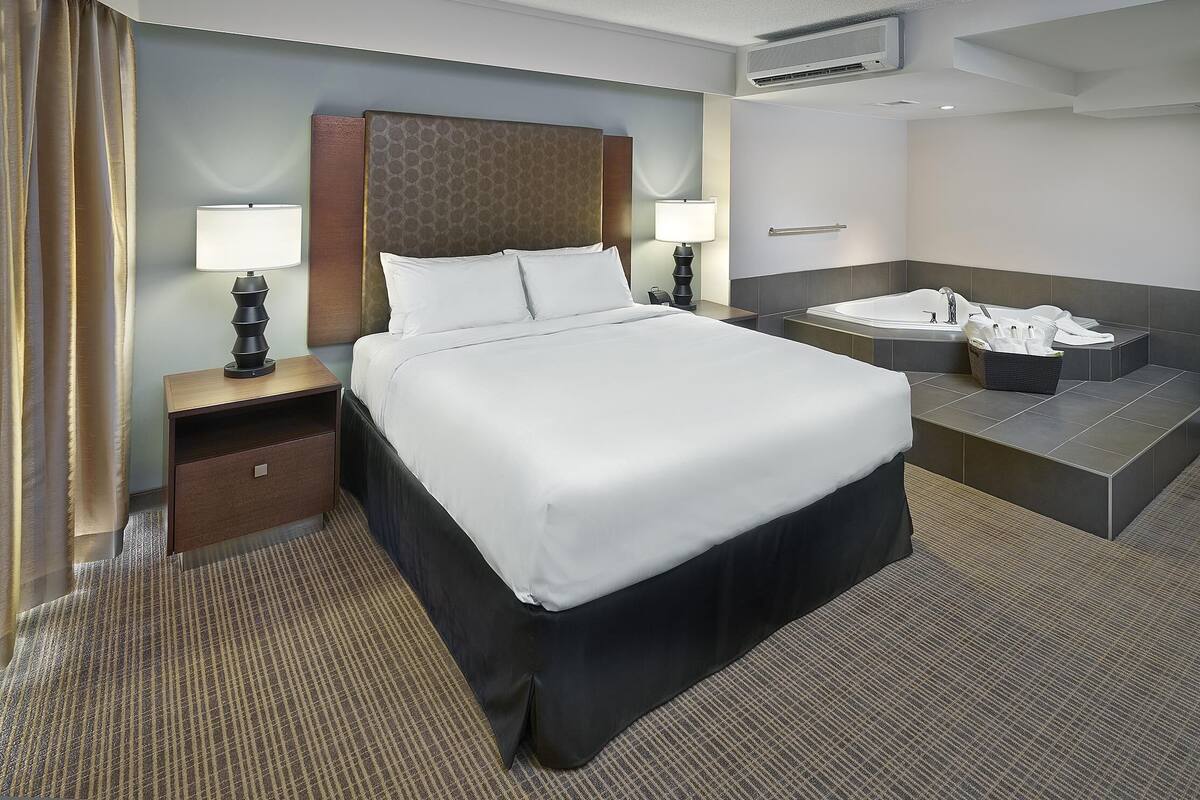 A hotel room with a large bed, two bedside tables with lamps, a small desk, a bathroom with a bathtub, and a shower.