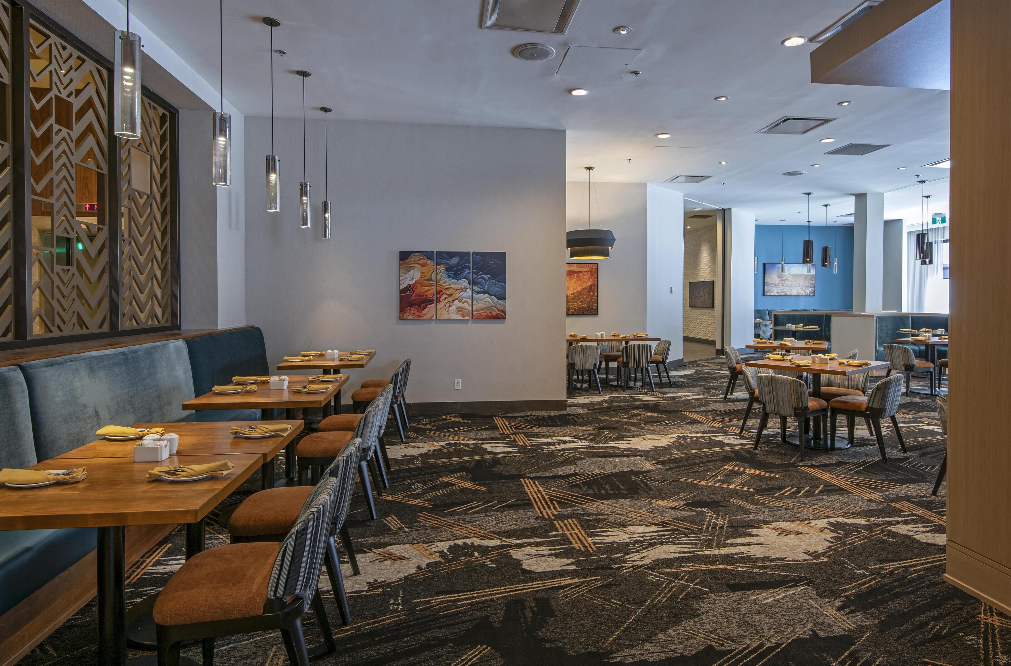 A hotel dining area with booths and tables, patterned carpet, pendant lighting, and artwork on the walls.