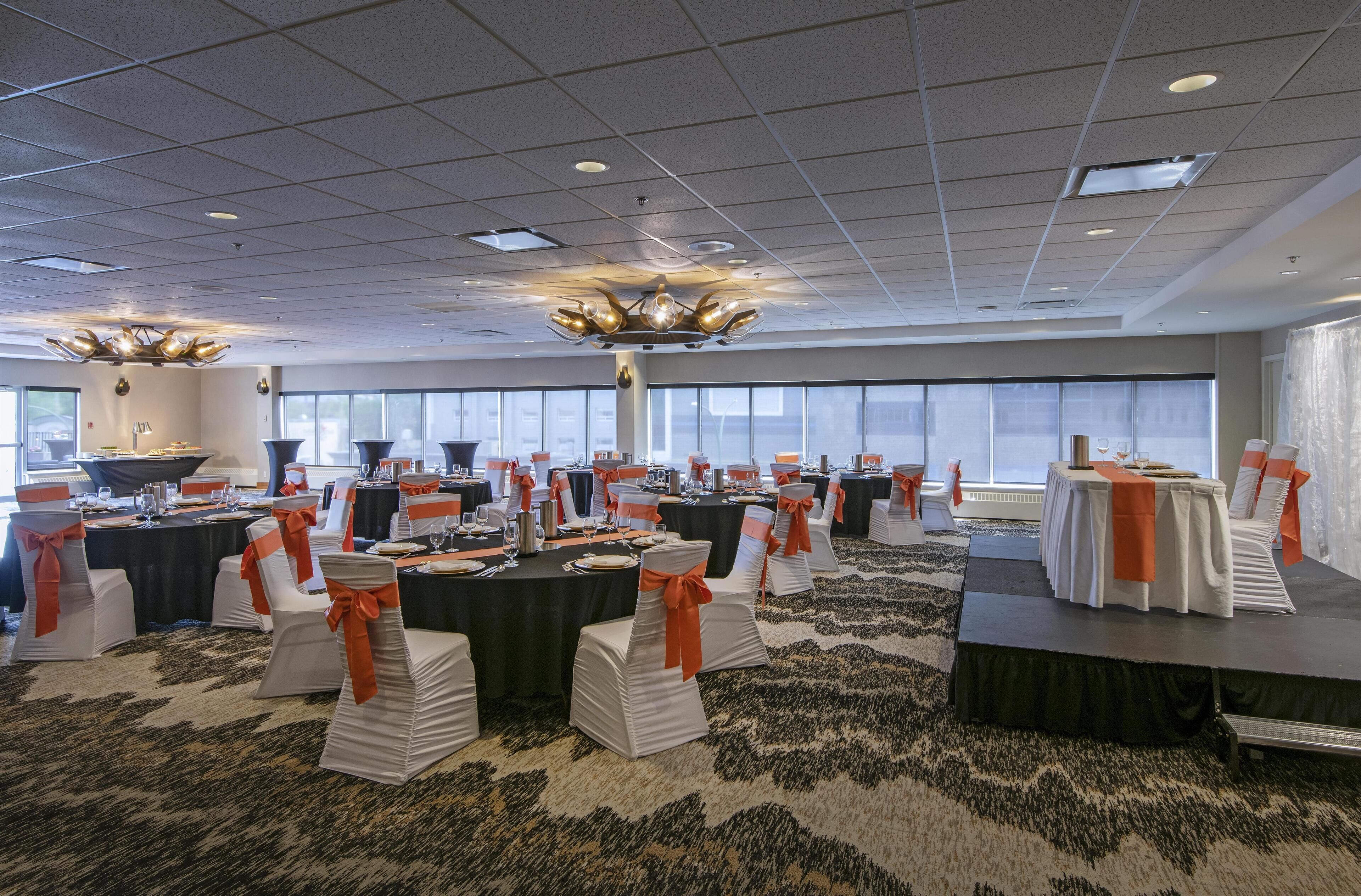 A banquet hall with tables set for a formal event, featuring white chairs with orange sashes, a stage, and large windows.