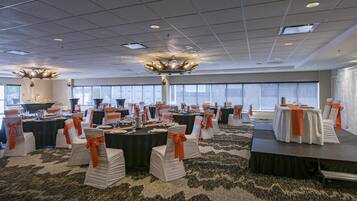 A banquet hall with tables set for a formal event, featuring white chairs with orange sashes, a stage, and large windows.
