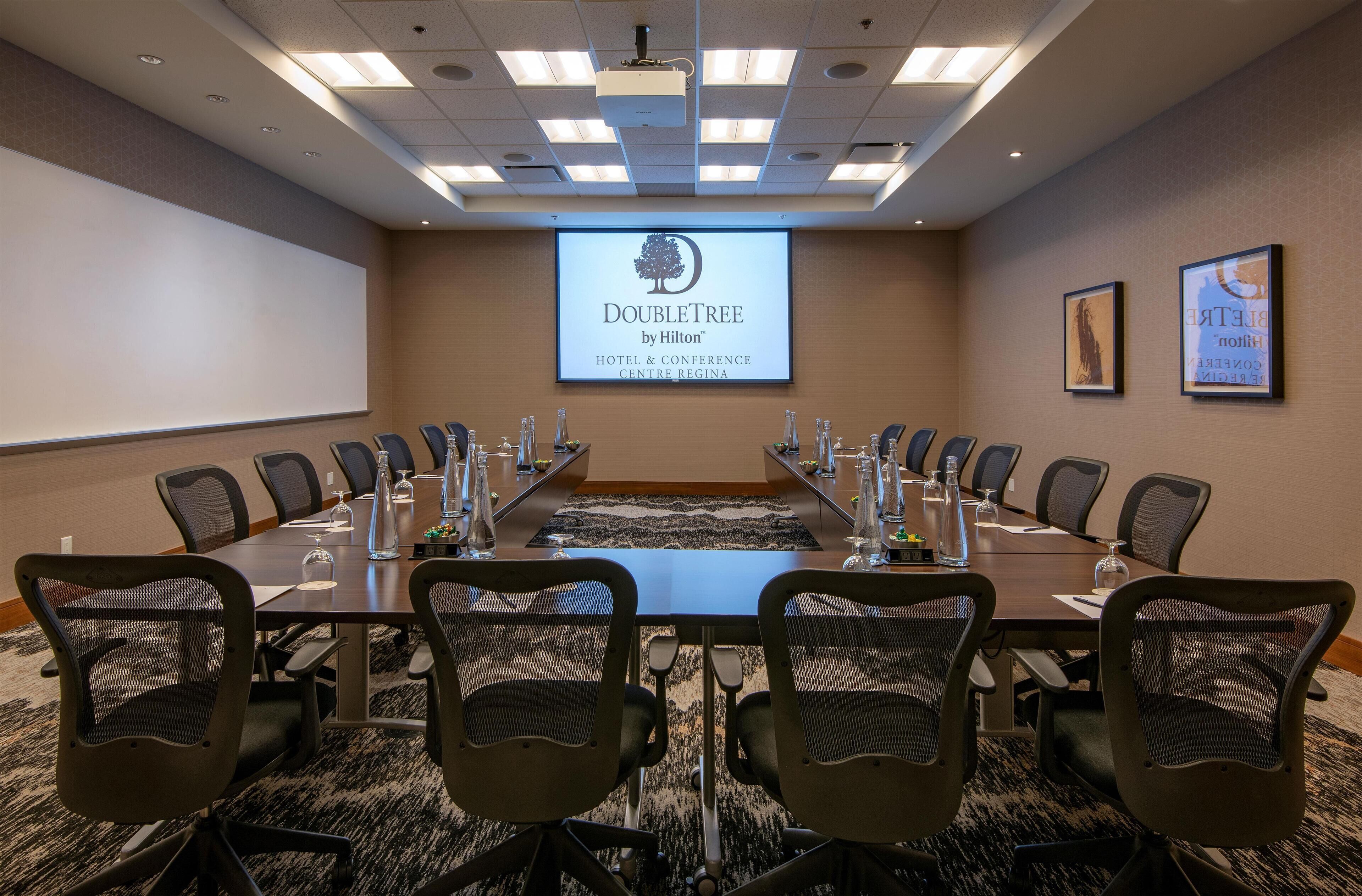 Ein Konferenzraum mit einem langen Tisch, Stühlen, einer Projektionsleinwand und Teppichboden.