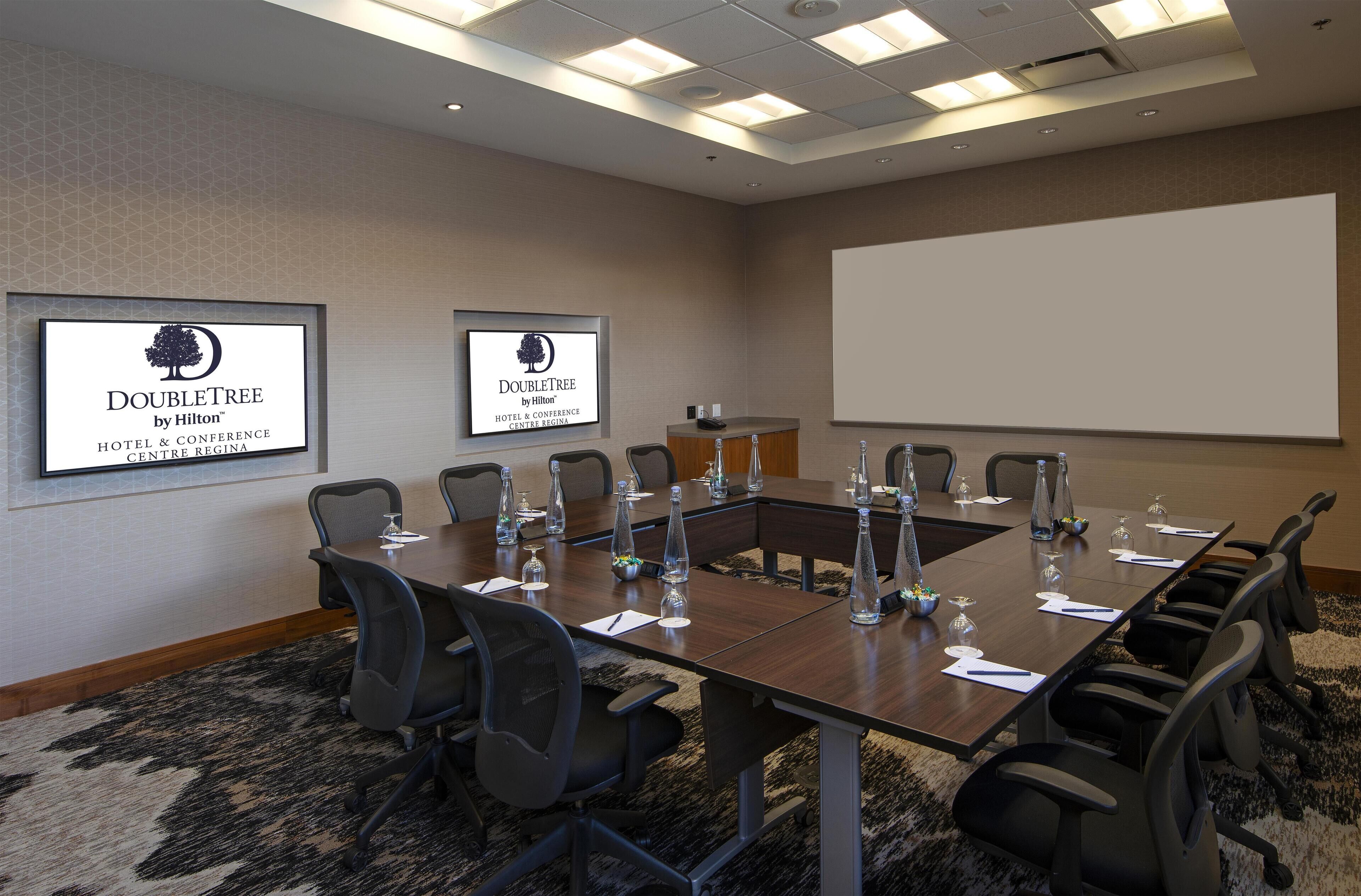 A conference room with a long table, chairs, a projector screen, and two monitors displaying the DoubleTree by Hilton logo.