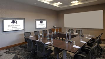A conference room with a long table, chairs, a projector screen, and two monitors displaying the DoubleTree by Hilton logo.