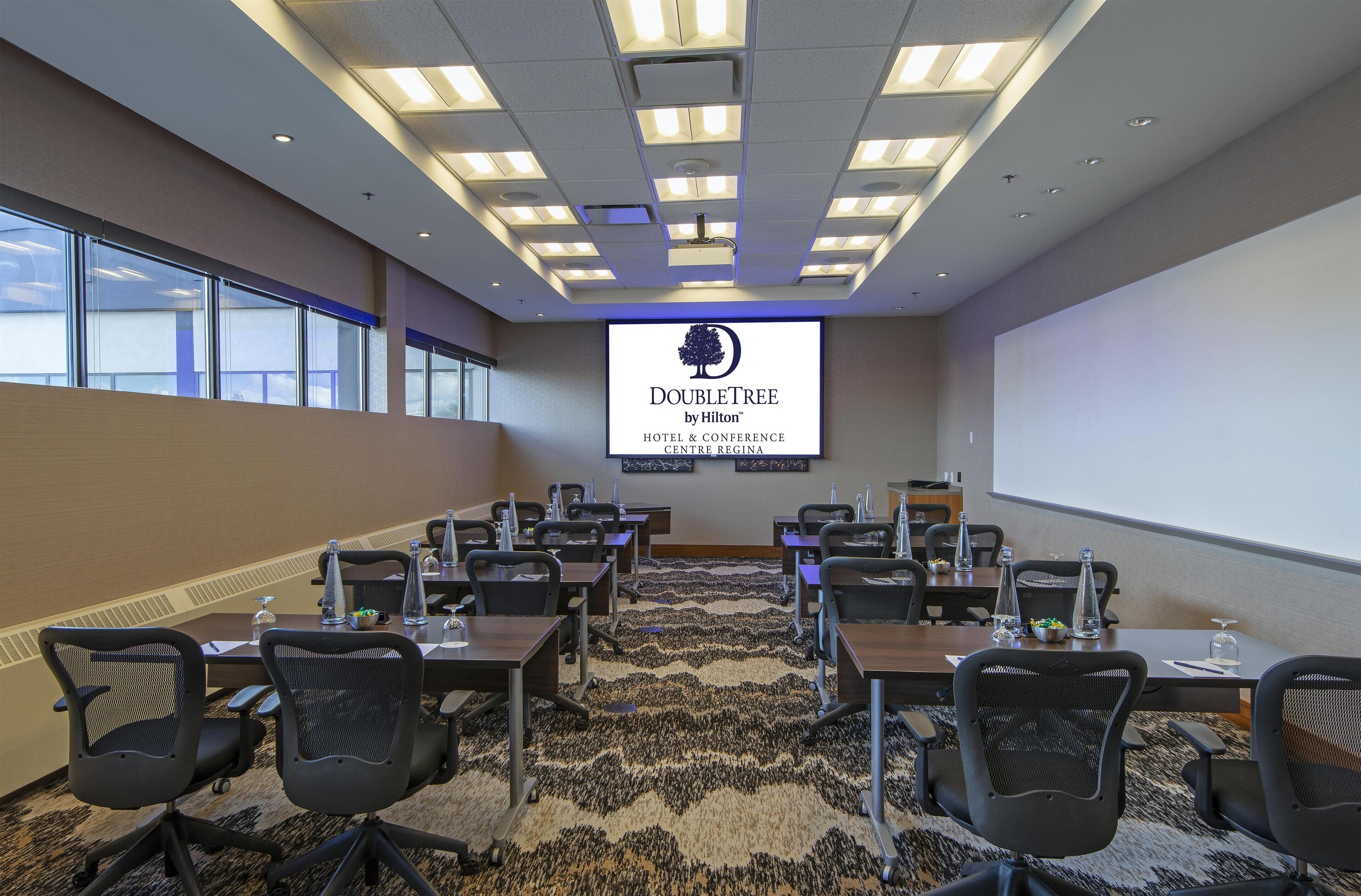 A conference room with a large screen displaying the DoubleTree by Hilton logo, multiple tables with chairs, and a carpeted floor.