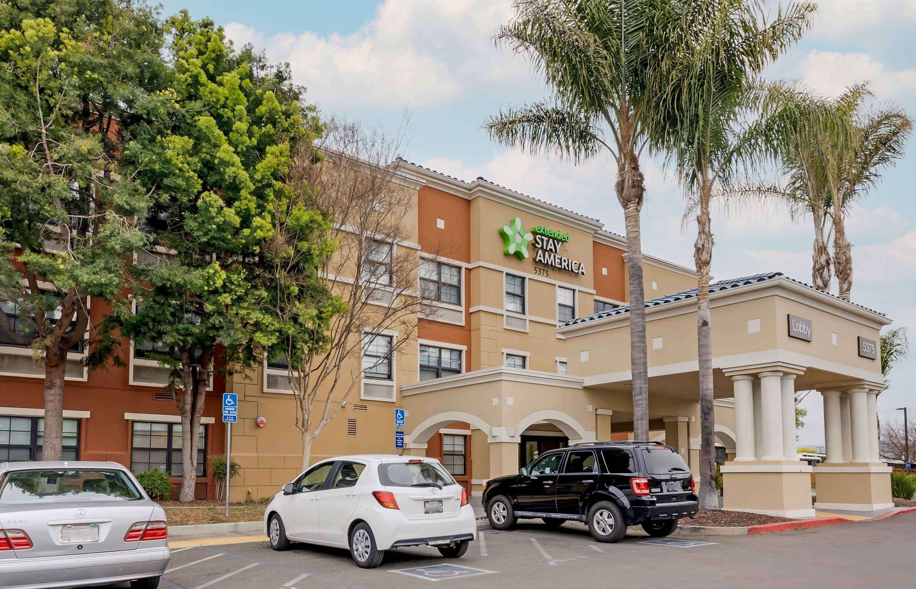 A Stay America hotel with a beige facade, palm trees, and a parking lot with cars.